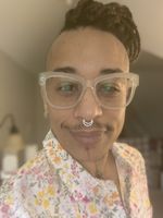 Close up face shot of a light-skinned person with a light mustache wearing clear glasses, a white septum ring, and braids on the top of their hair. The sides of their head are shaved close. They have a face tattoo of a red line down their chin, extending to a black line with a spider in their neck dip AKA jugular notch. They are wearing a buttoned-collar white shirt with pink, yellow and orange small flowers. The background is blurry.