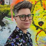 A nonbinary trans person, white with salt and pepper short hair, gently smiles into the camera. They have black glasses and a brightly patterned collared shirt, standing against a bright yellow spray painted backdrop.