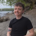 Eli Haan faces the camera and smiles while at a lake beach with water, sand, trees and wood debris in the background. Eli has fair skin, short, medium brown hair, and color arm tattoos. They are photographed from the torso and above, and is wearing a black t shirt.