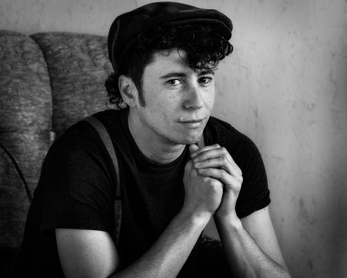 Vintage style black-and-white photograph of Kai Tillman posing with bent arms on an armchair. Kai has fair skin and dark, curly ear-length hair with sideburns. He is wearing a dark flat cap, a dark t shirt and vintage patterned suspenders.