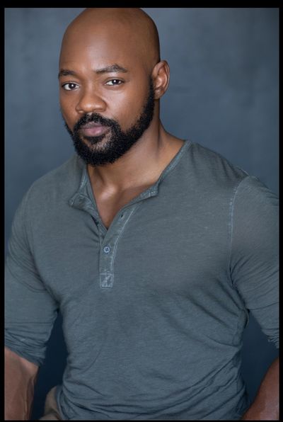 Professional studio portrait of Brian Michael Smith from the torso and above. Brian has dark skin, dark facial hair and a bald hairstyle. He is wearing a earthy blue coloured long sleeve shirt with his forearms exposed.