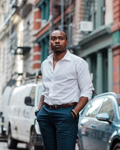 Professional portrait of Tiq Milan standing on a sophisticated city street that is softly blurred behind him. Tiq has dark skin, a very short dark hairstyle, and light facial hair. He is styled in dark blue tailored pants, a belt, and white button down shirt with arms rolled up to reveal a watch and bracelet.