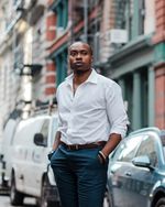 Professional portrait of Tiq Milan standing on a sophisticated city street that is softly blurred behind him. Tiq has dark skin, a very short dark hairstyle, and light facial hair. He is styled in dark blue tailored pants, a belt, and white button down shirt with arms rolled up to reveal a watch and bracelet.