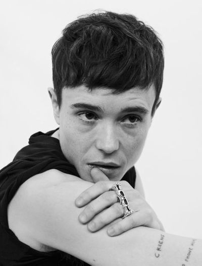 Black-and-white closeup of Elliot Page. Elliot is a nonbinary trans man with light skin and short dark hair. He wears a black tank top and a set of brass knuckles, with his right shoulder turned towards camera and his left hand resting on his arm, gently thumbing his lip as he stares contemplatively past the camera.