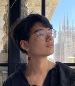 Mira De Koven seen in profile before a window to European-looking buildings, a poetic expression on their face. Mira is a nonbinary and mixed race person with short dark hair, big round glasses, and a glass cherry earring.