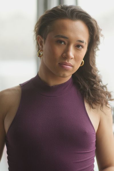 Chicahua Zipactonal Martinez standing near a window with soft, diffused daytime lighting. Chicahua has tan skin, brown eyes and brown shoulder-length hair. They are wearing a purple sleeveless turtleneck paired with gold earrings.