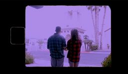 Kai & Love together in Love's imagination. Both wear brightly-colored plaid shirts and face away from camera, towards a white building surrounded by palm trees. There is a purple haze over the image, and the colors of the surroundings are washed out by bright daylight.