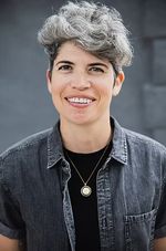 Brooke Stern Sebold faces the camera with an open smile. They have tall wavy grey hair, light skin, and dark eyes. They wear a denim button-up, black t-shirt and pendant necklace.