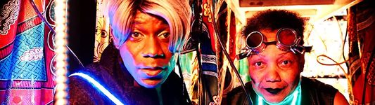 Two characters from "Ancestors, Can You Read Us? (Dispatches from the Future)" stare directly at the camera. The person on the left (played by Ravyn Wings) has a blonde bob, medium brown skin, and wears a black jacket with sash made of LED lights, while the person on the right (played by Gloria Swain) is slightly shorter with a short blow-out, light brown skin and round cyberpunk glasses. They appear to be in a tent made of kitenge or other African-style batik with some hanging wires.