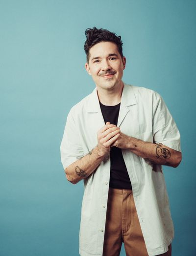 Amos Mac stands in front of a dark eggshell-blue background and stares into the camera, his fist in his hand. He has light skin, a mustache, and a stylish wave of dark hair atop his head. He wears a white labcoat over a plain black shirt and chestnut-brown jeans.