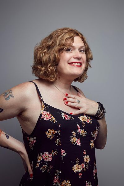 Studio portrait of Lilly Wachowski smiling and holding her hand to her chest in front of a grey background. Lilly is a white trans woman with curly shoulder-length strawberry blonde hair and tattooed arms. She wears red lipstick, red nail polish and a black dress with flower patterns.