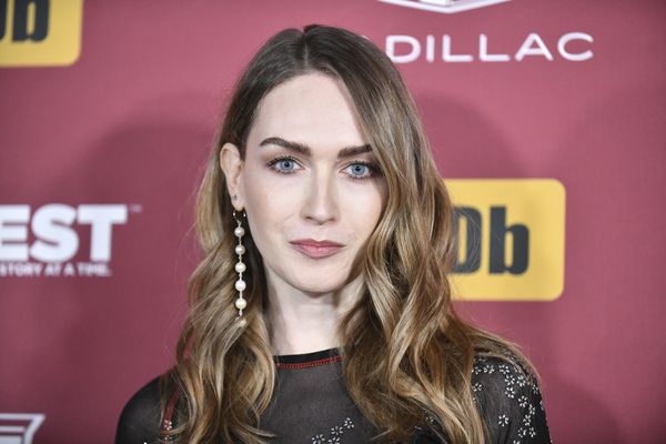 Red carpet headshot of Jamie Clayton with raised eyebrows and a faint smile. Jamie is a white trans woman with dirty blonde hair and blue eyes. She wears a dangling string of pearls as an earring and a dark nylon dress accentuated with clusters of little metal flower petals.