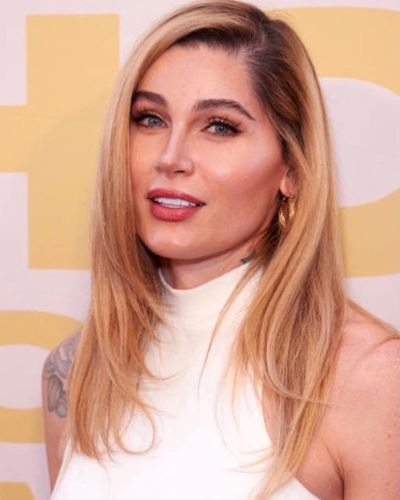 Red carpet headshot of Trace Lysette with a slight smile. Trace Lysette is a light-skinned trans woman with blond hair past her shoulders. She wears a sleeveless white top and has tattoos on her arms.
