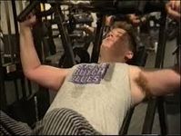 Still of a person weightlighting, likely Leslie Feinberg. Ze has an expression of concentraion, light skin, short blond hair, and wears a grey shirt that says "Stone Butch Blues".