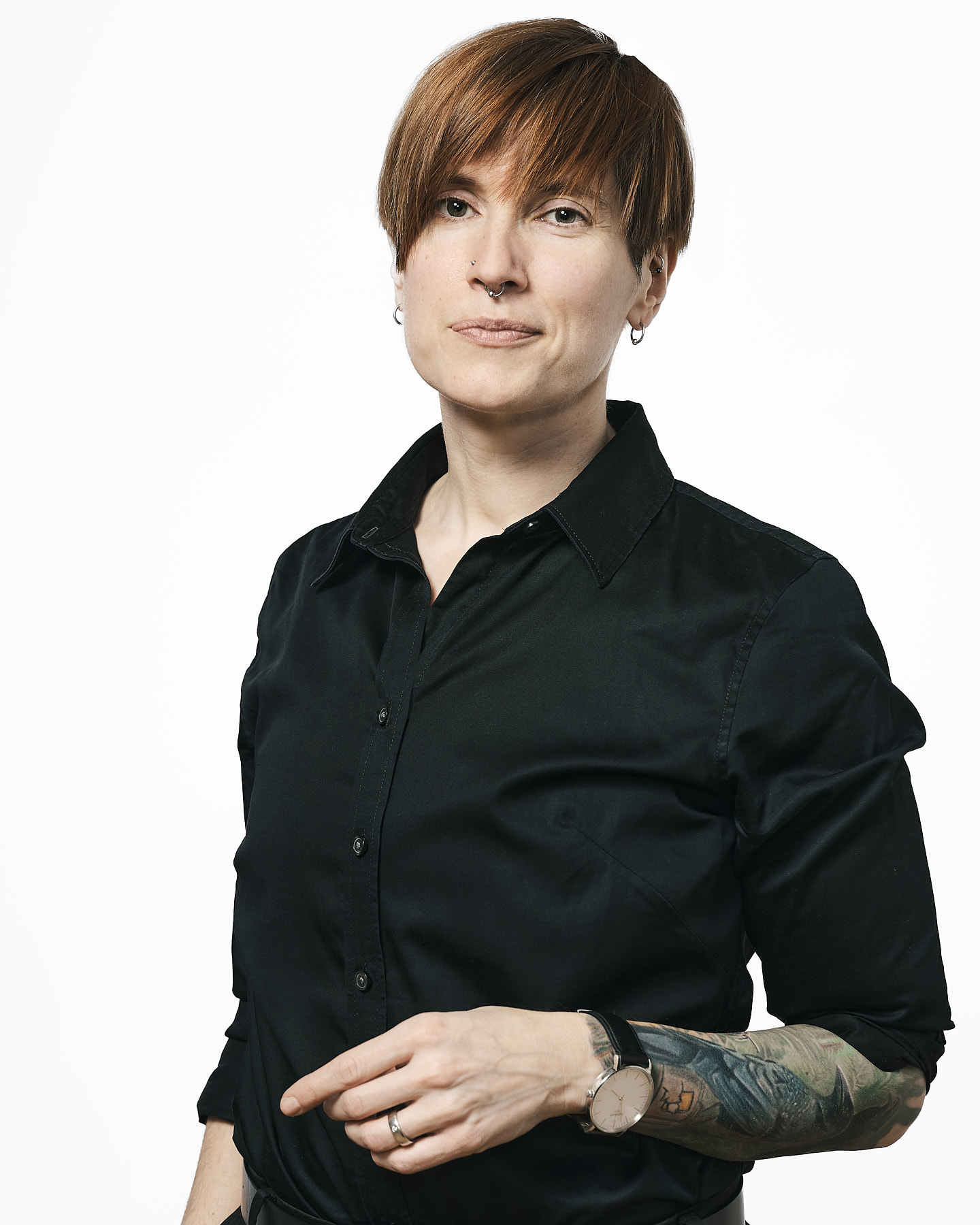 Constance has light skin, short red hair, and wears a button-down shirt with sleeves rolled to the elbows. They stand against a white background, facing the camera and smiling slightly.