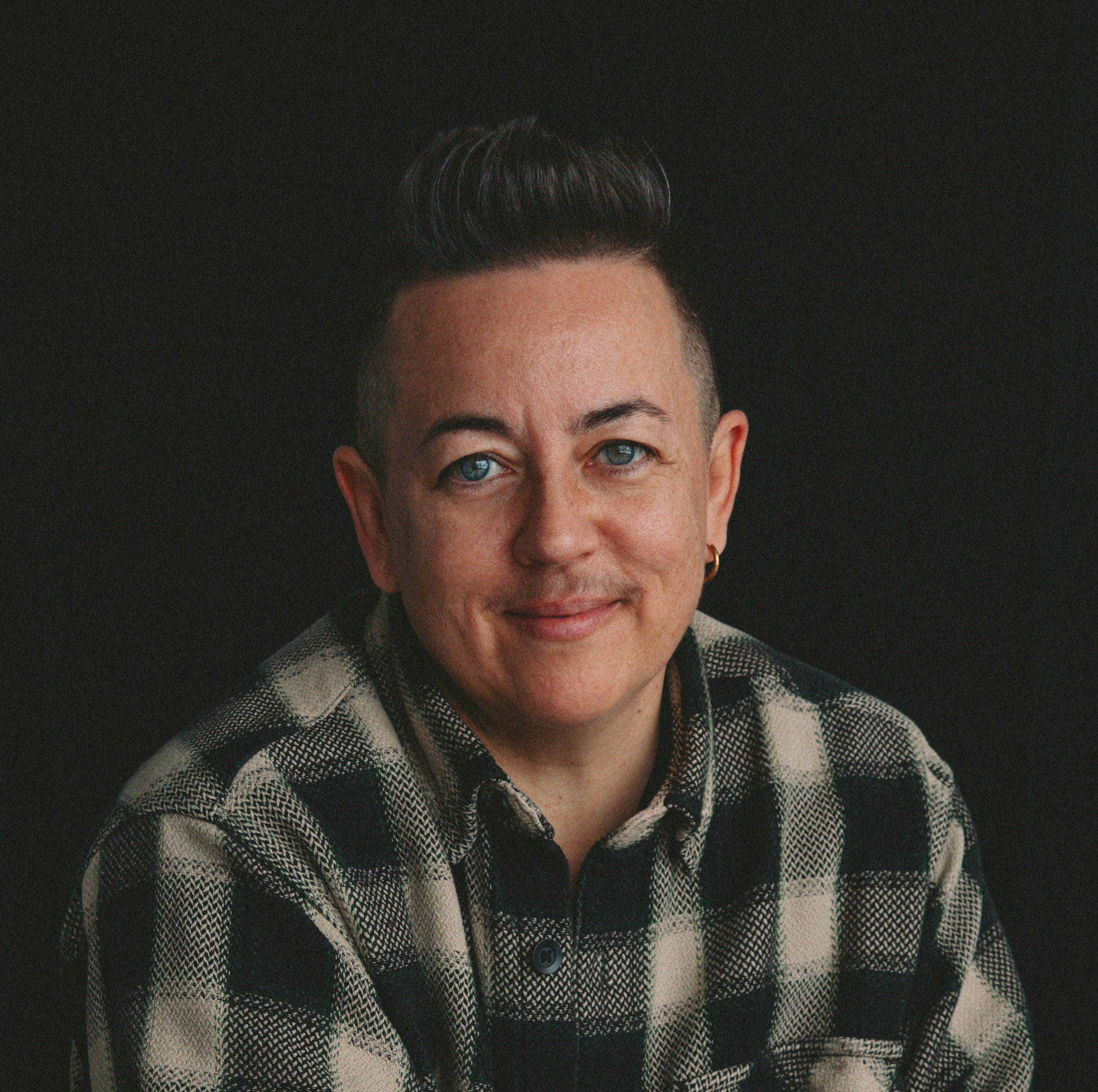 A smiling headshot of Evie Johnny, who has light skin, blue eyes, and dark hair cropped short on the sides. They wear a black and beige plaid button-up shirt and a gold hoop in their left ear.