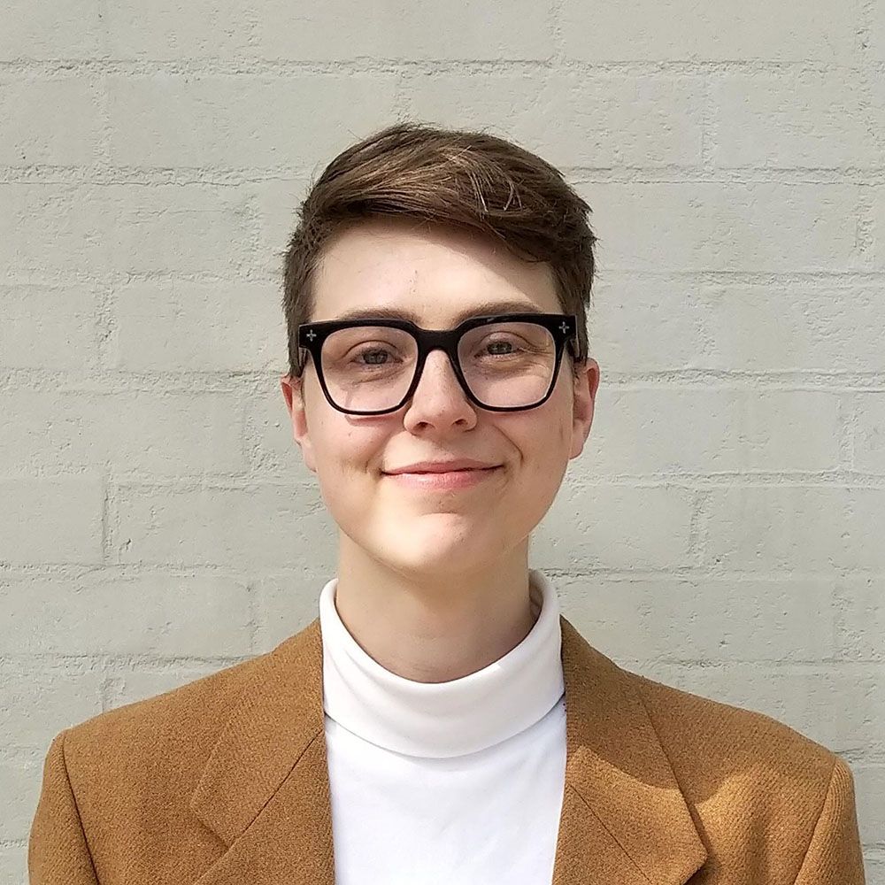 Orvis has light skin and short brown hair. They wear glasses, a white turtleneck, and a cognac-coloured blazer. They stand in front of a white wall smiling at the camera.