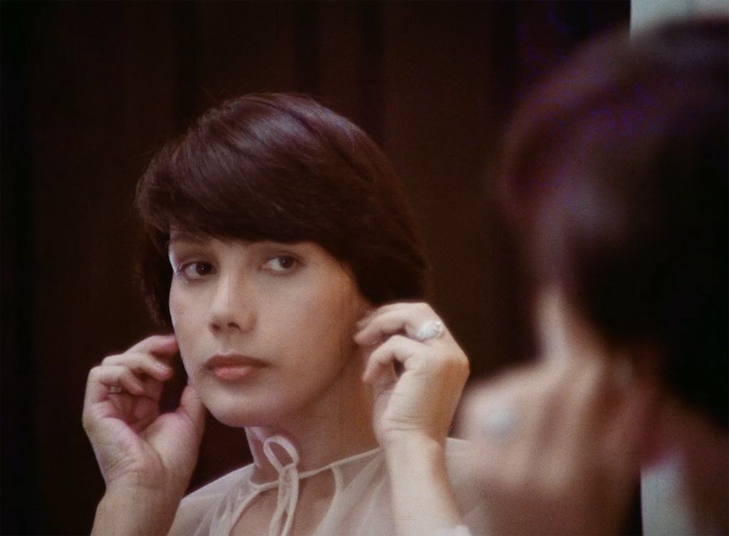 Still from Let Me Die a Woman (1977). A light-skinned character with short brown hair looks in the mirror and fixes their earrings.They are looking in the mirror and the camera is focused on their reflection in the mirror with the person slightly off to the side blurred out. They are wearing a white blouse and have a silver ring on their ring finger.