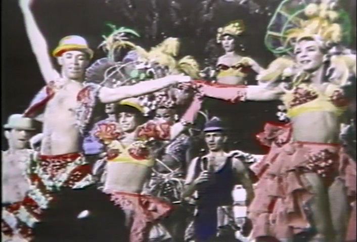 Still from Stormé: The Lady of the Jewel Box (1987). A group dances in fancy parade costumes. Two people in the front are the focal point. They are holding hands. The person on the right has a yellow and red bra and a red skirt with red gloves on. The person on the left has a red and white vest and matching pants but no t-shirt underneath and a yellow and red top hat with their left arm raised in the air.