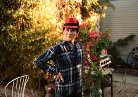 Hida Viloria is a Latine person with brown eyes, short dark hair and light skin. They are wearing a dark blue and white plaid shirt and red fedora with a black strap. They stand with their hands on their hips and slight smile on a leafy patio.