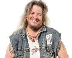 Alec Butler has long sandy hair, a beard, light skin, and a big smile. They are wearing a denim vest with patches with a white tank top underneath, a leather necklace with a pendant, and have some tattoos on their arms and chest and piercings.