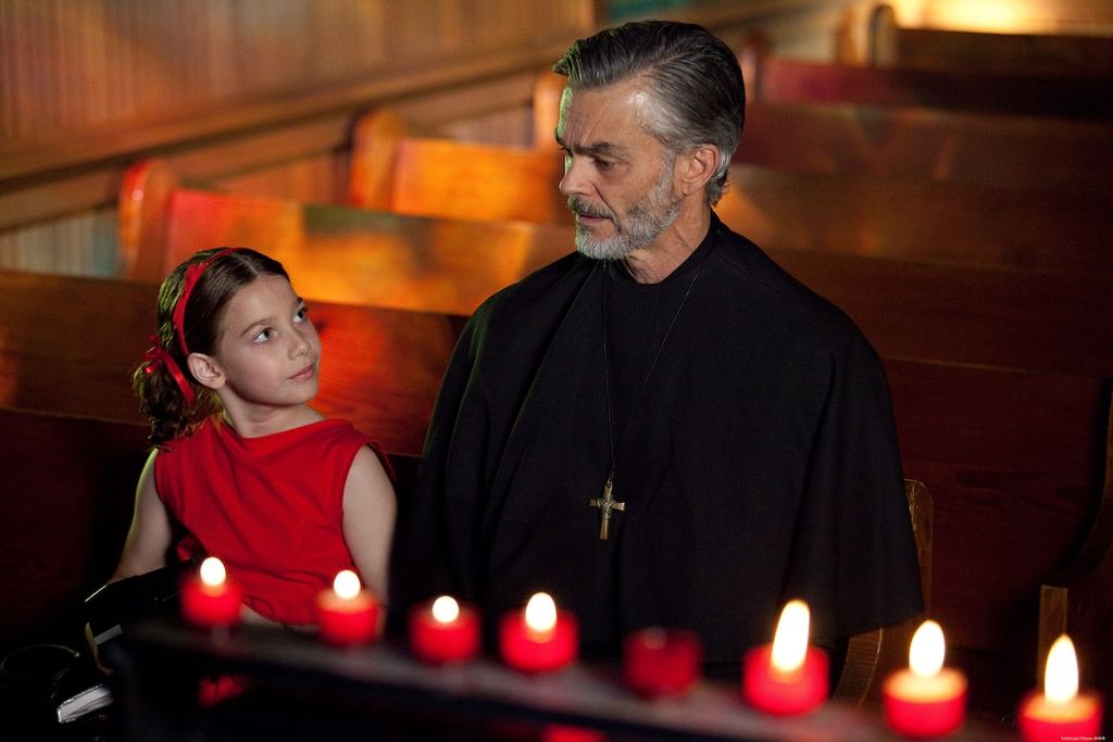 Still from Limina (2017). Alessandra, a gender-fluid child in a red dress with red bows in their hair, exchanges a curious glance with a priest sitting next to them in a pew. Red votive handles frame the shot. The background is a row of slightly blurred brown wooden pews.