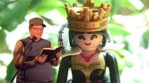 Still from Just Dandy (2013). Filmmaker Theo Jean Cuthand stands in front of a microphone and reads from a book. A plastic figurine of a smiling queen is projected behind him. The smiling queen has a gold crown, a red necklace, and a gold and black dress. Theo is wearing a grey tank top, a light brown brimmed hat, and wearing glasses.